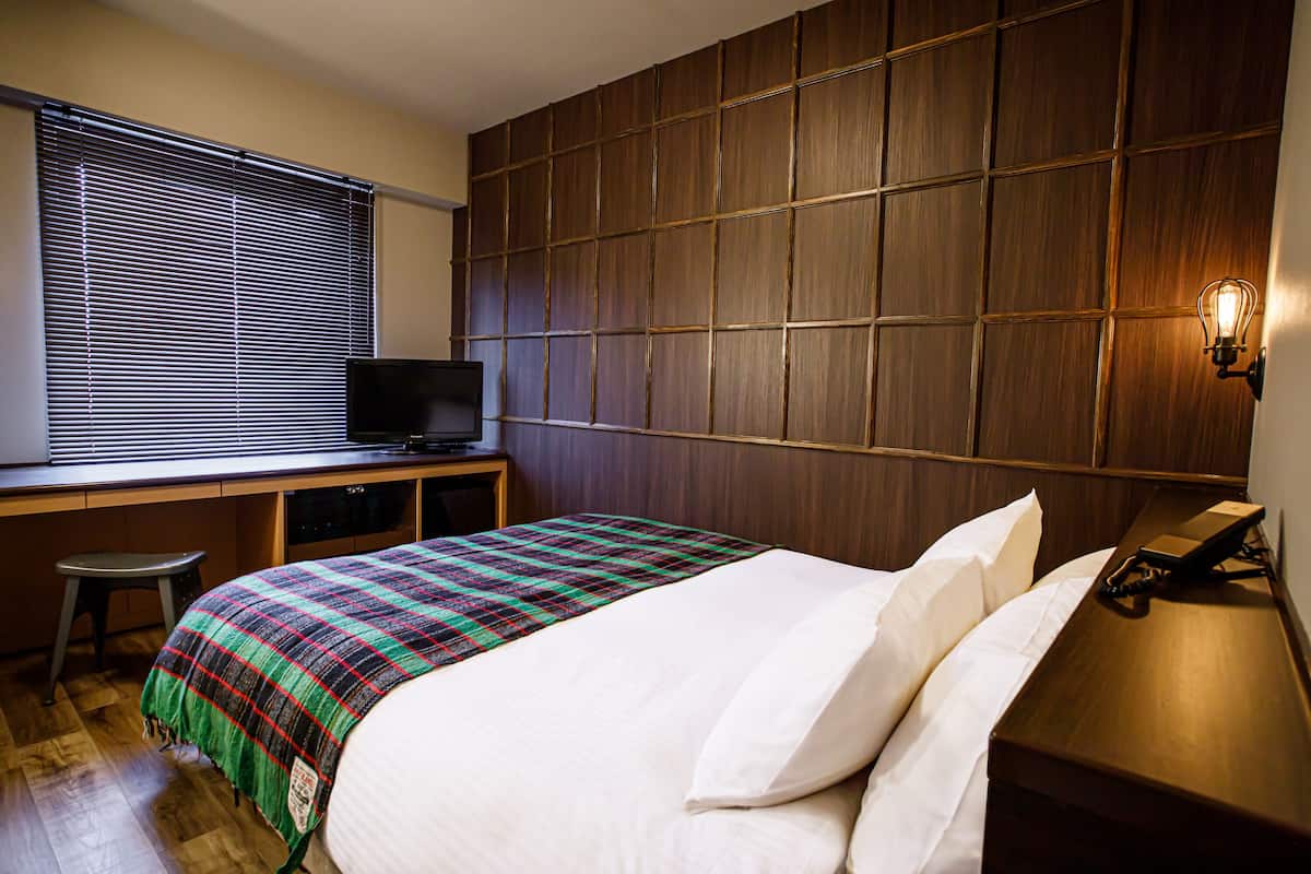 A hotel room with a bed, a desk with a television, a stool, and a wall-mounted light.