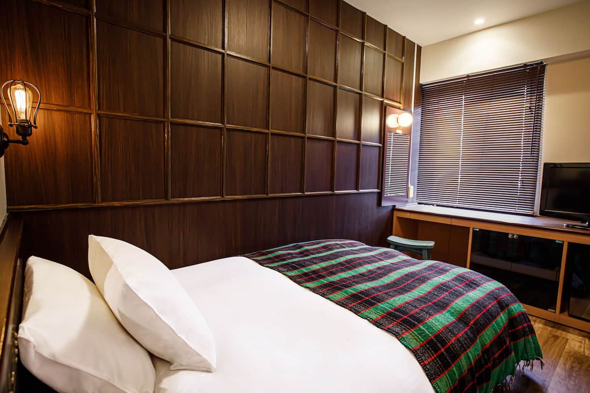 A hotel room with a bed, a TV, and a window with blinds.