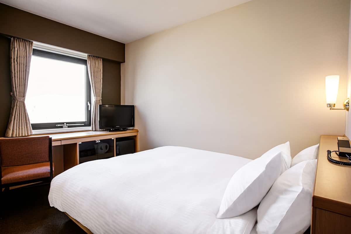 A hotel room with a bed, a desk with a television, a chair, and a bedside table with a lamp.
