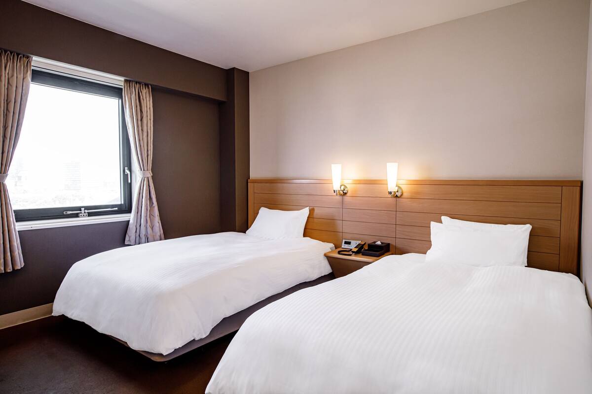 A hotel room with two beds, a window with curtains, bedside lamps, and a wooden headboard.