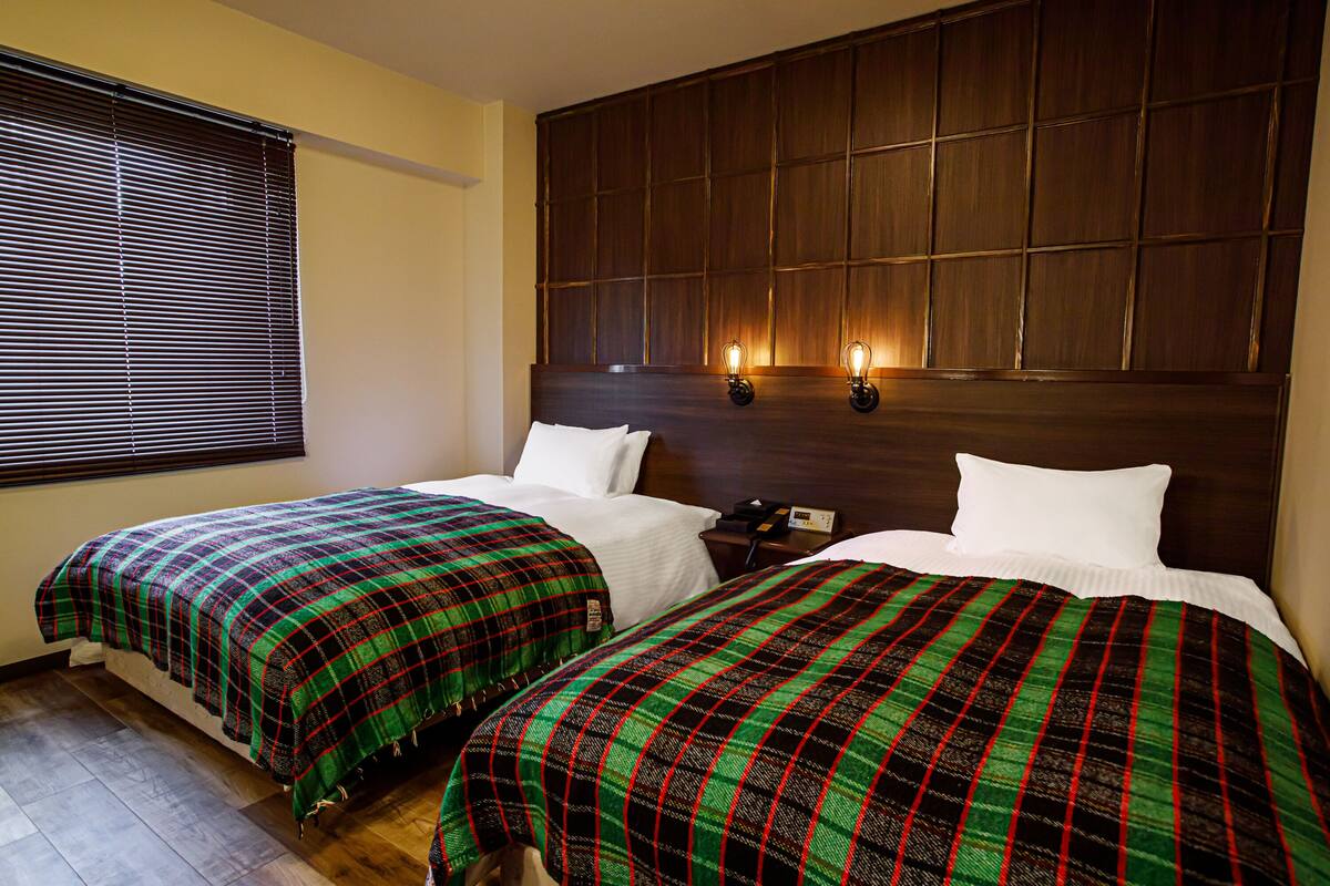 A hotel room with two beds, each with a plaid bedspread, a wooden headboard, and two wall-mounted lamps.