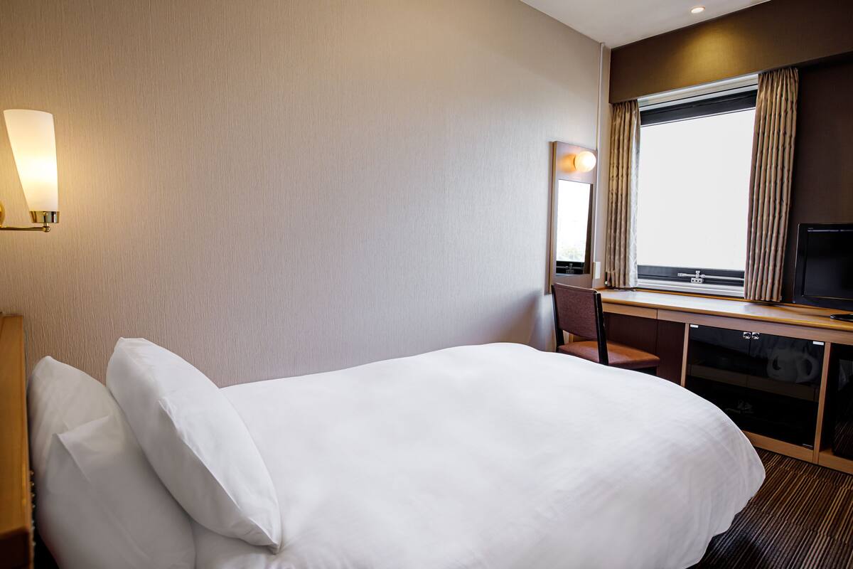 A hotel room with a bed, a desk with a chair, a television, and a window with curtains.