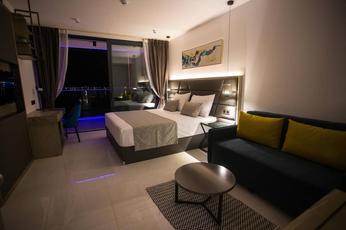 Ein modernes Hotelzimmer mit Bett, Sofa, kleinem Tisch und Blick auf die beleuchtete Stadt bei Nacht.