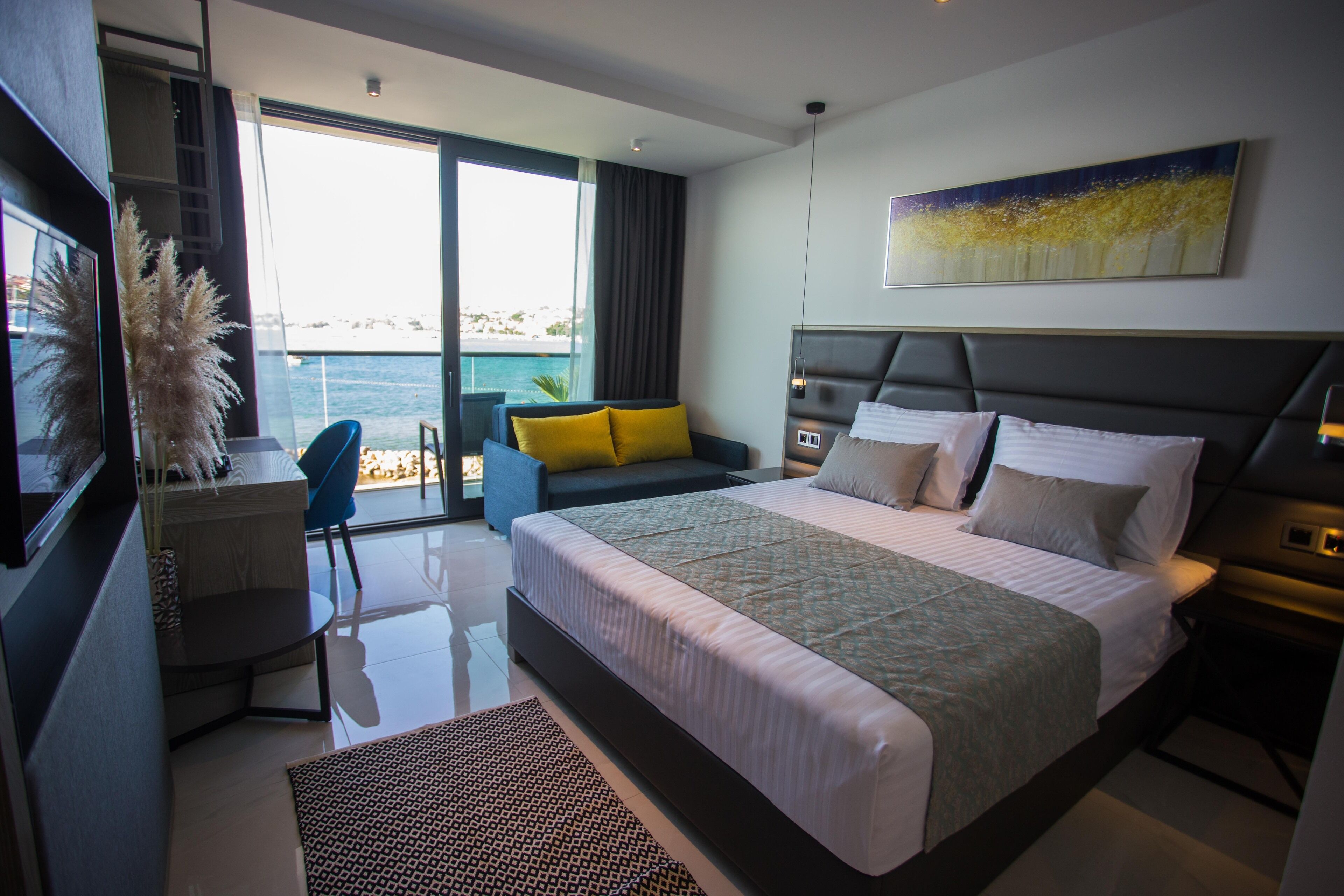 A modern hotel room with a large bed, a desk, a chair, and a view of the sea.