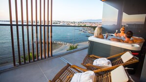 Un balcone con vista sul mare e sulla città, una vasca idromassaggio con persone e poltrone prendisole in legno.