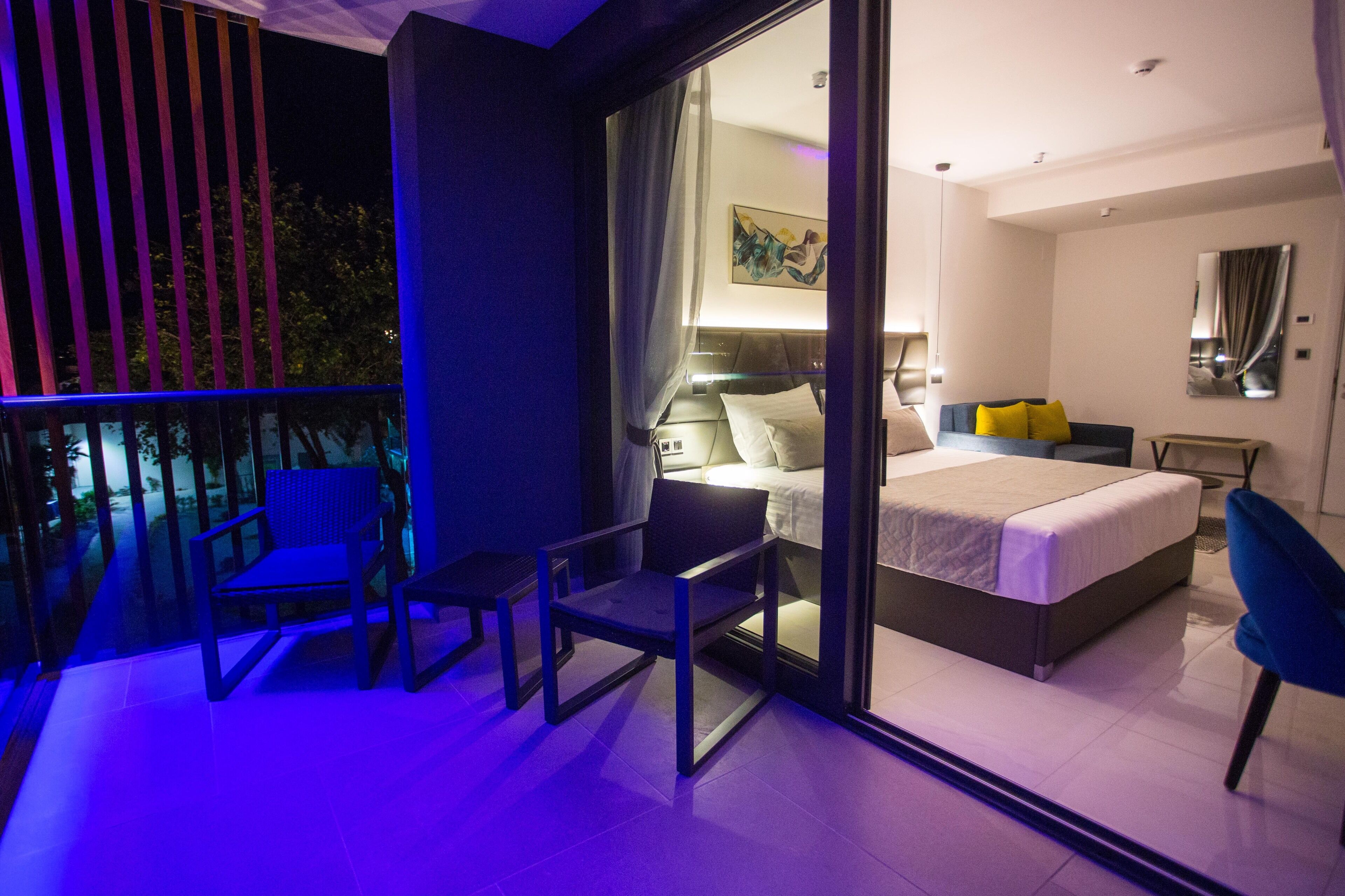 A modern bedroom with a large bed, a desk, and a chair. There is a balcony with a view of trees.