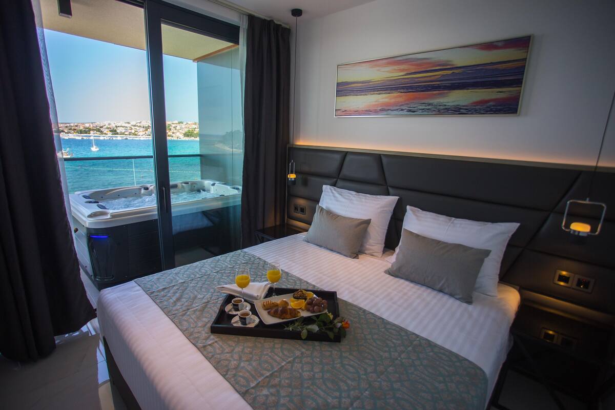 Ein Hotelzimmer mit Bett, einem Frühstücksbrett und Meerblick mit Booten. Ein Bild hängt an der Wand.