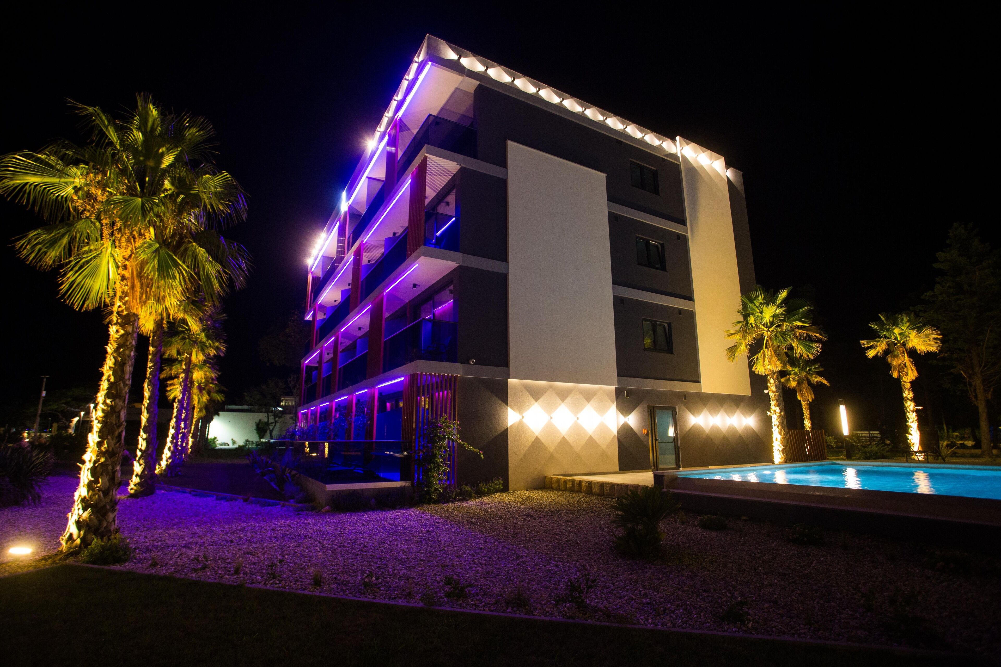 Un hotel de varios pisos con piscina, palmeras e iluminación exterior.