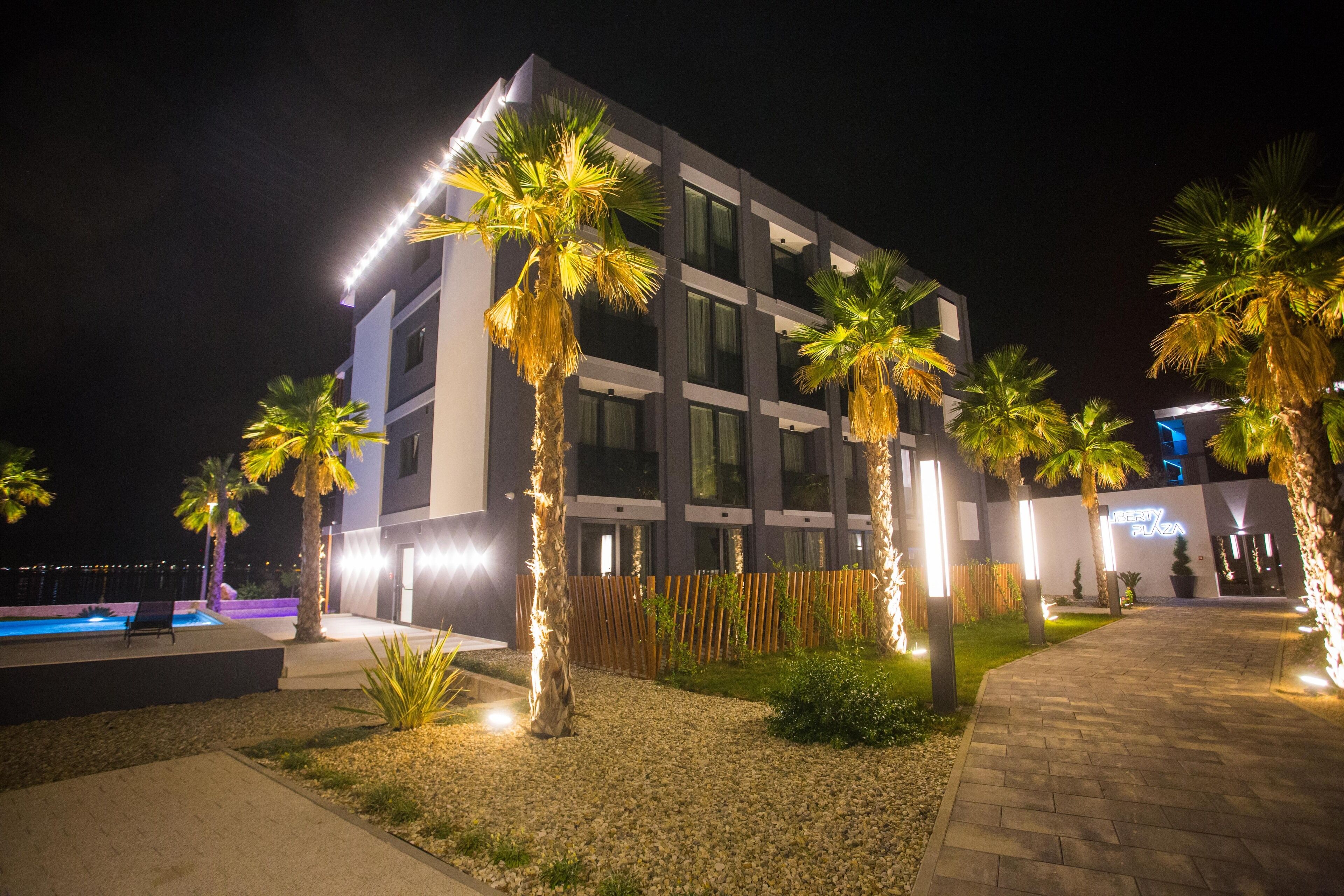 A modern building with a swimming pool, palm trees, and a well-lit entrance.