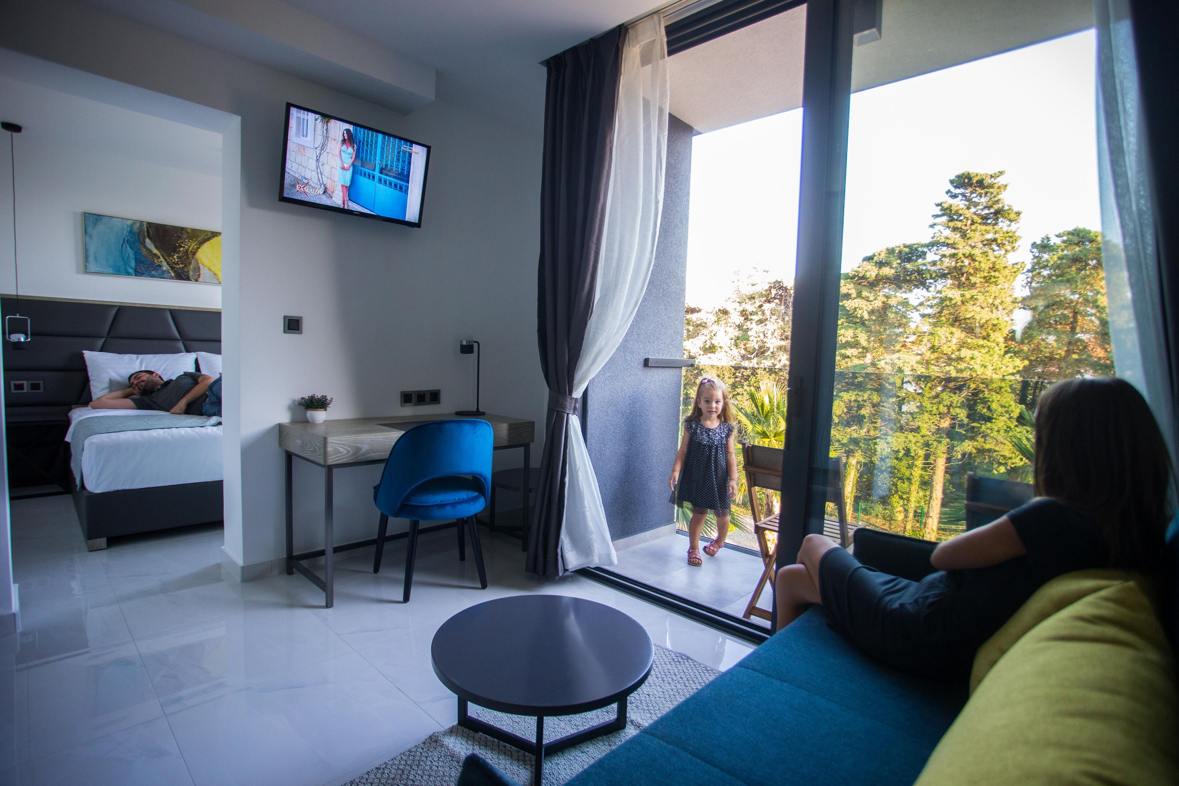 Quarto de hotel moderno com cama, escrivaninha, televisão e varanda com vista para árvores.