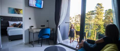 Quarto de hotel moderno com cama, escrivaninha, televisão e varanda com vista para árvores.