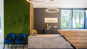 Een receptie met een groene wand, twee blauwe stoelen, een houten balie en een groot raam met uitzicht op bomen.