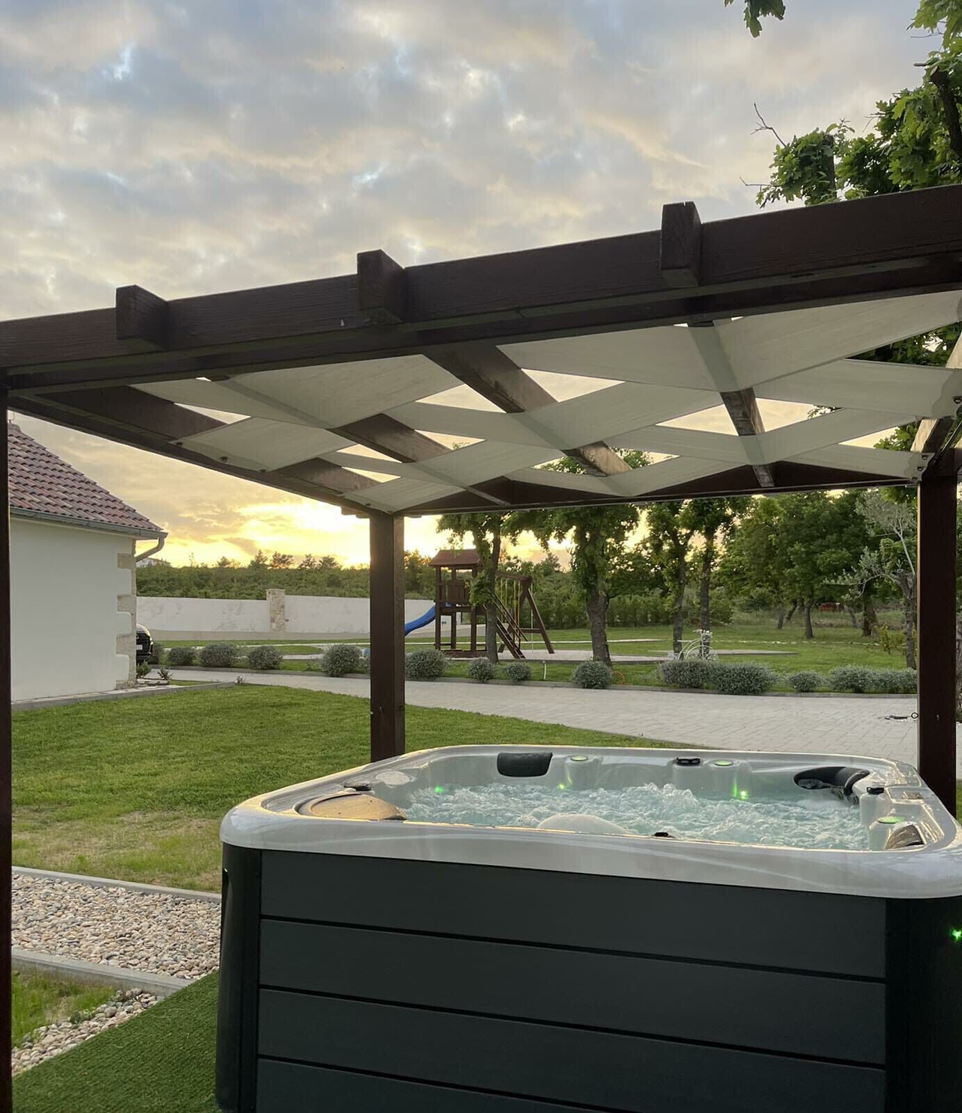 A hot tub under a wooden pergola in a backyard with a playground in the background.