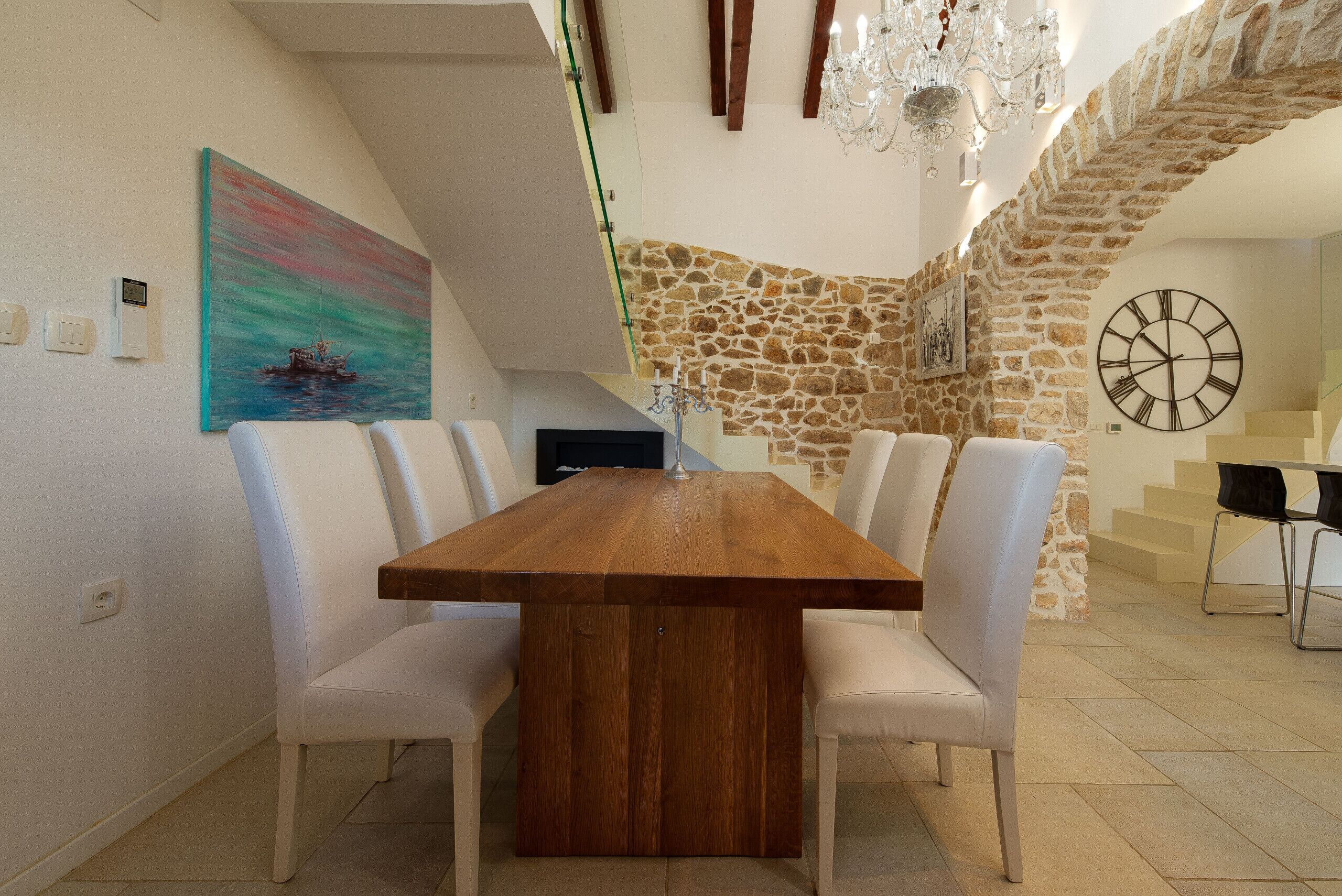 A dining area with a wooden table and white chairs, a painting on the wall, a clock, and a stone wall.