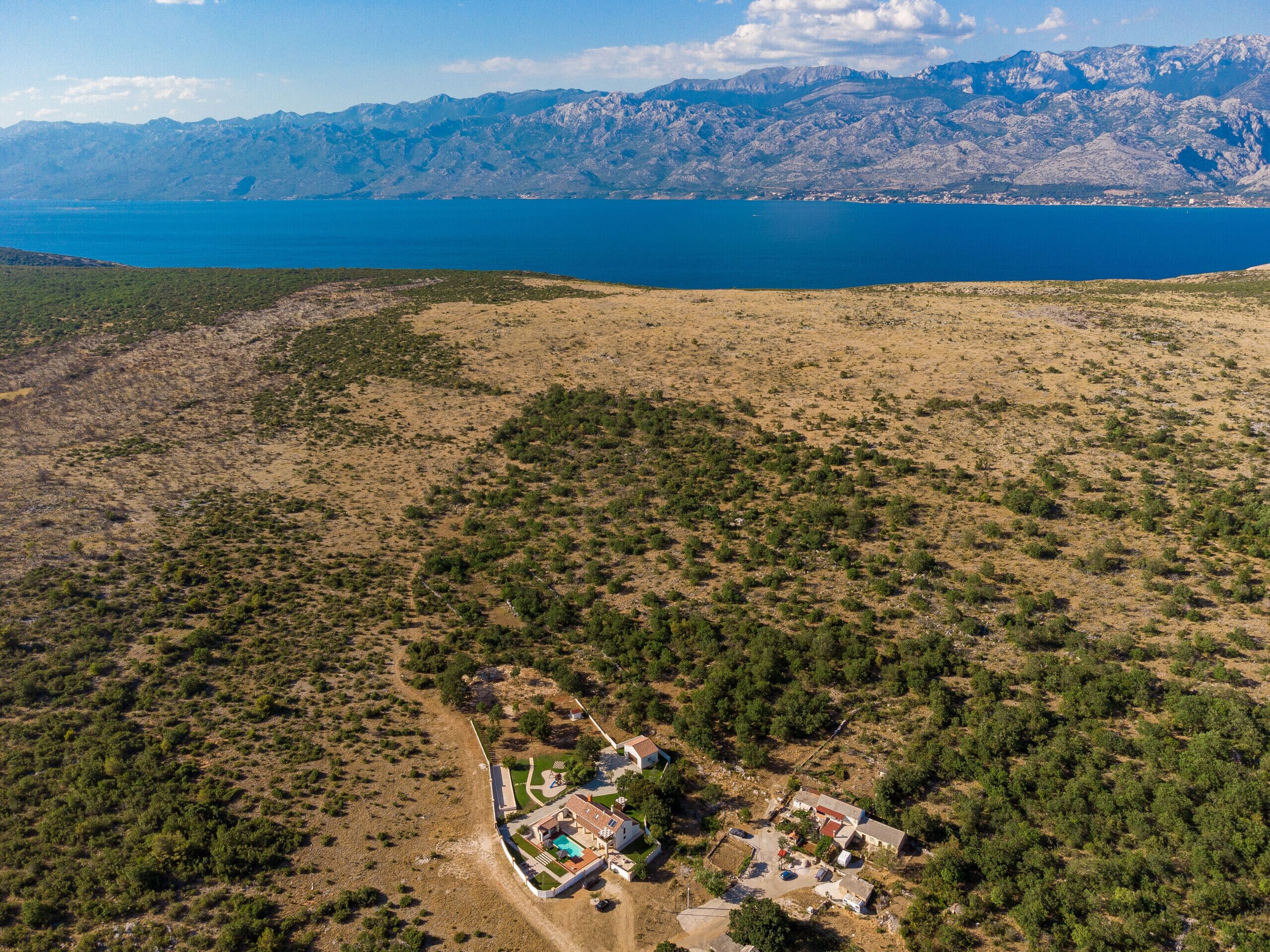 An aerial view of a property with a swimming pool, a tennis court, and a small cluster of buildings surrounded by sparse vegetation and mountains in the background.