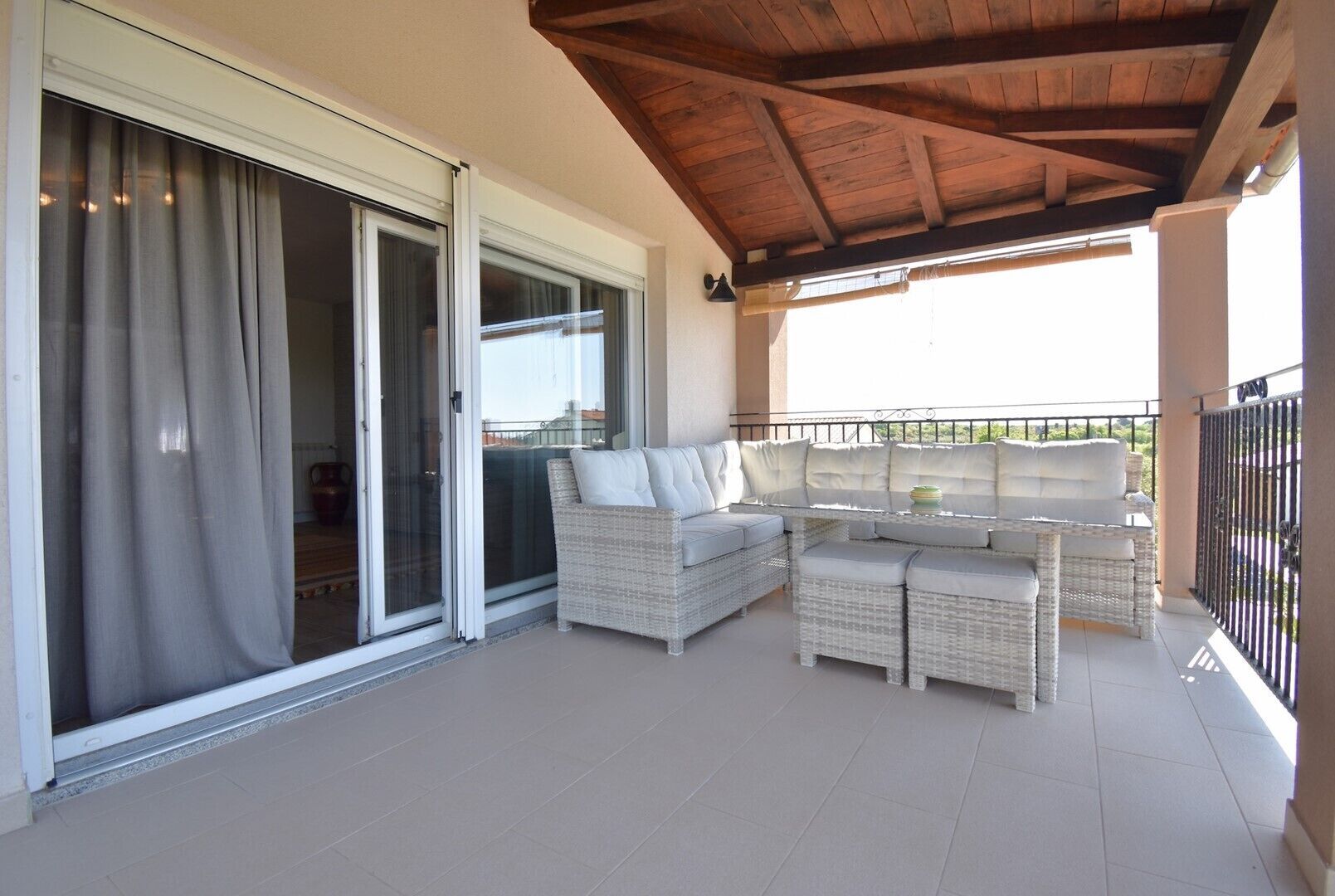 A covered balcony with a wicker sofa and table, sliding glass doors, and a wooden ceiling.