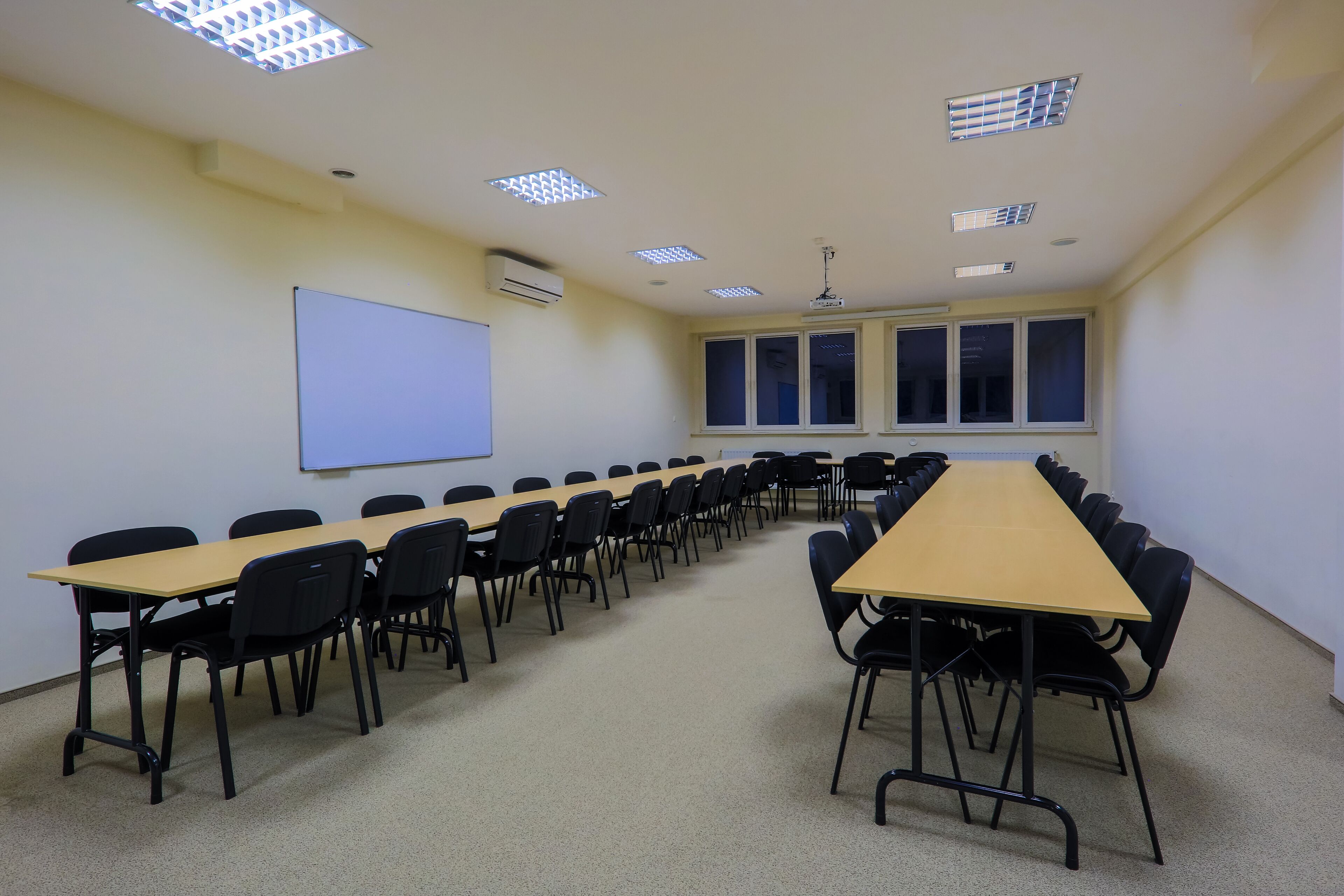Un salón de conferencias con una mesa larga, sillas negras, una pantalla de proyector y ventanas.
