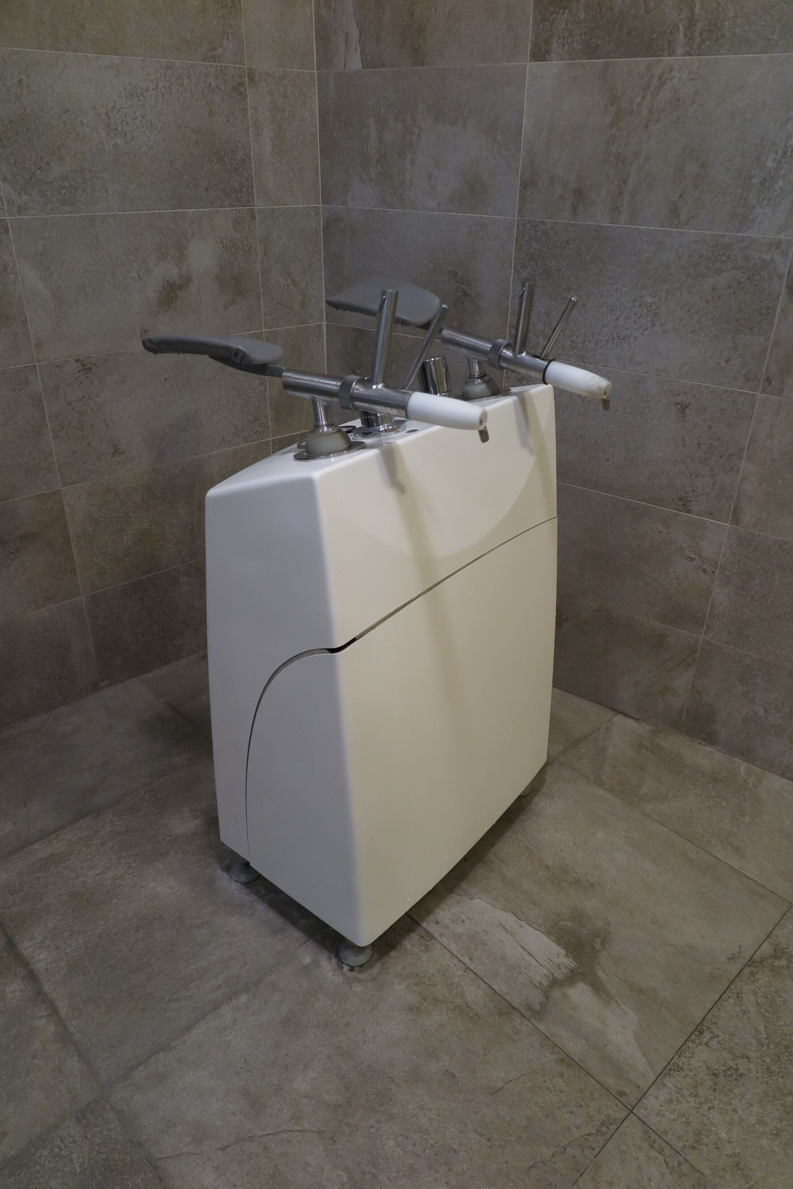 Una máquina de ejercicio blanca, estacionaria, con manijas y un pedal, ubicada frente a una pared con azulejos en una habitación con suelo de losa.