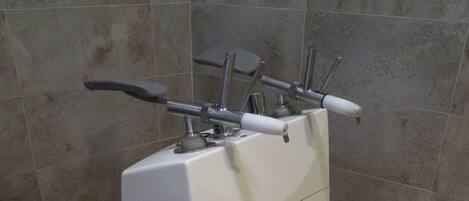 Una máquina de ejercicio blanca, estacionaria, con manijas y un pedal, ubicada frente a una pared con azulejos en una habitación con suelo de losa.