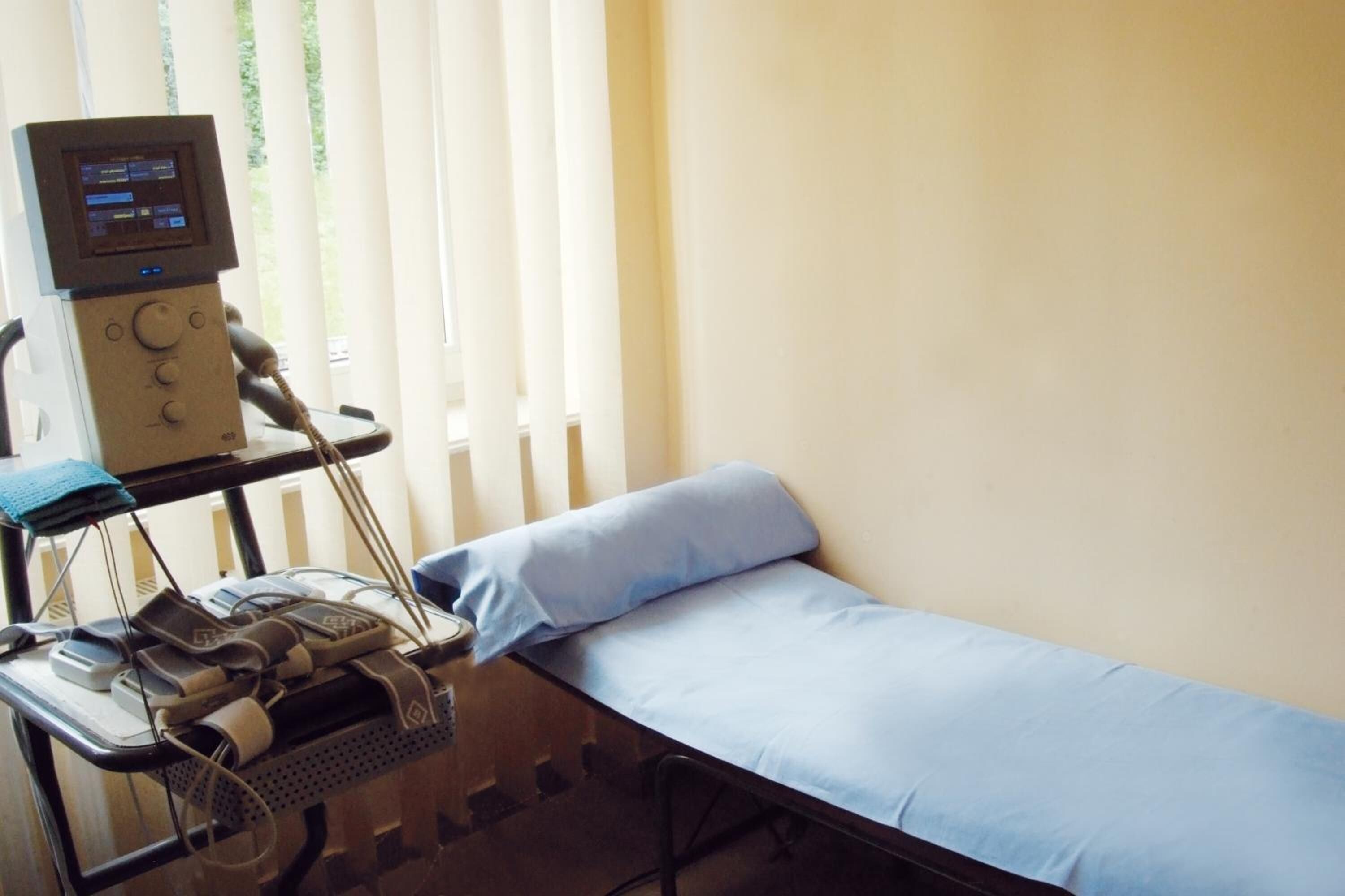A medical examination room with a bed, an examination trolley with medical equipment, and a window with blinds.