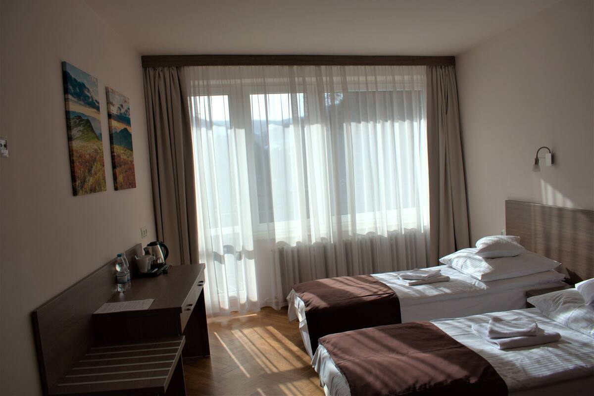 Ein Hotelzimmer mit zwei Betten, einem Schreibtisch und einem Fenster mit durchsichtigen Vorhängen.