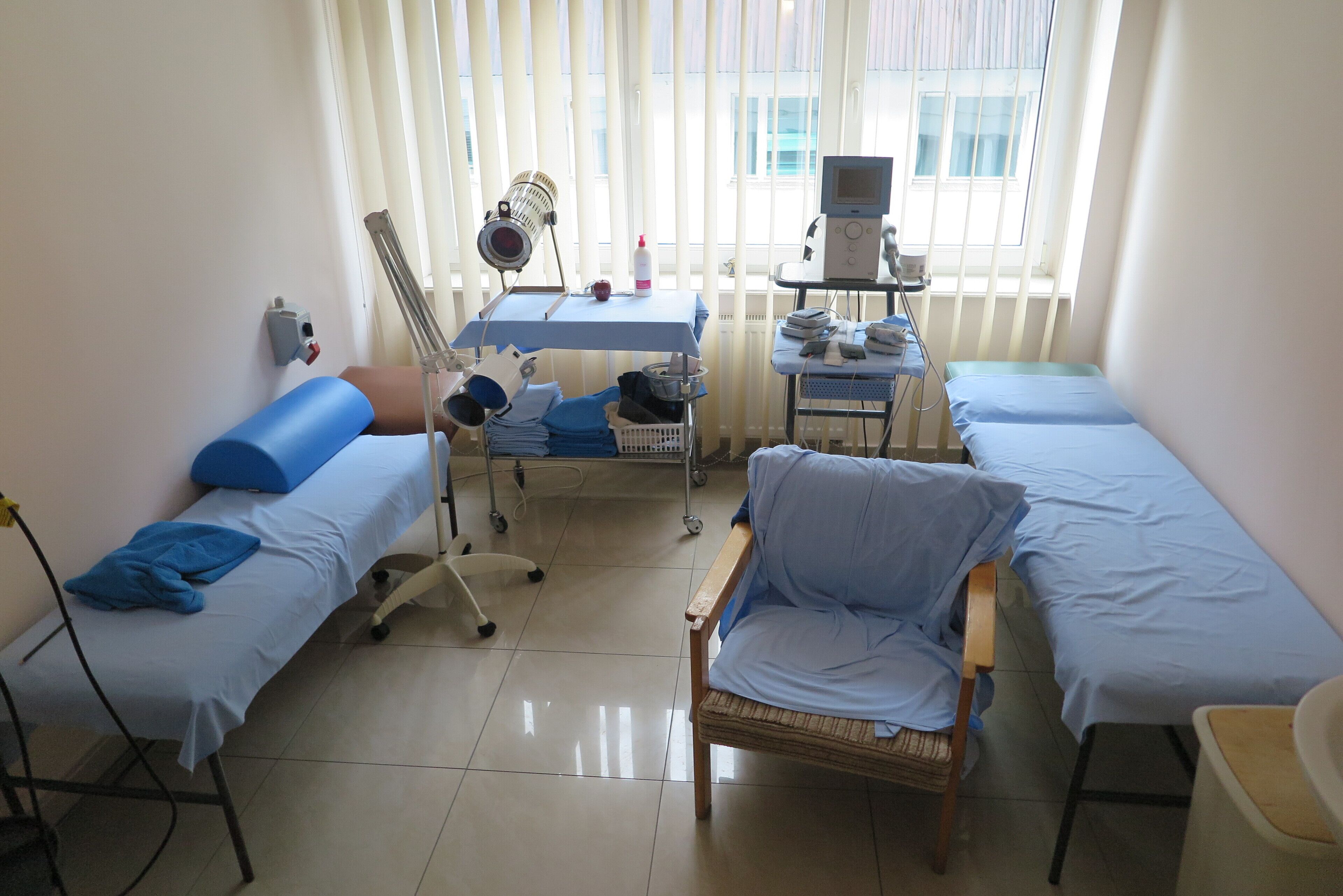 Une salle de consultation équipée de deux lits, d’une chaise, d’un chariot médical et d’une fenêtre avec des stores.