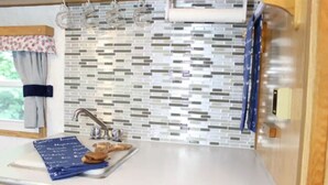 A kitchen with a tiled backsplash, a sink, and a window with curtains. - Nautical Cozy camper trailer on Lake Simcoe (Orillia)