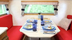 A dining area with a table set for four, red upholstered chairs, a window with curtains, and a view of greenery outside. - Nautical Cozy camper trailer on Lake Simcoe (Orillia)