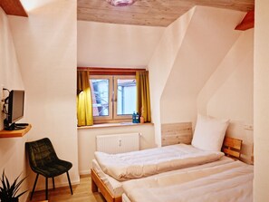 A small, cozy room with two beds, a desk with a computer, and a window with curtains.