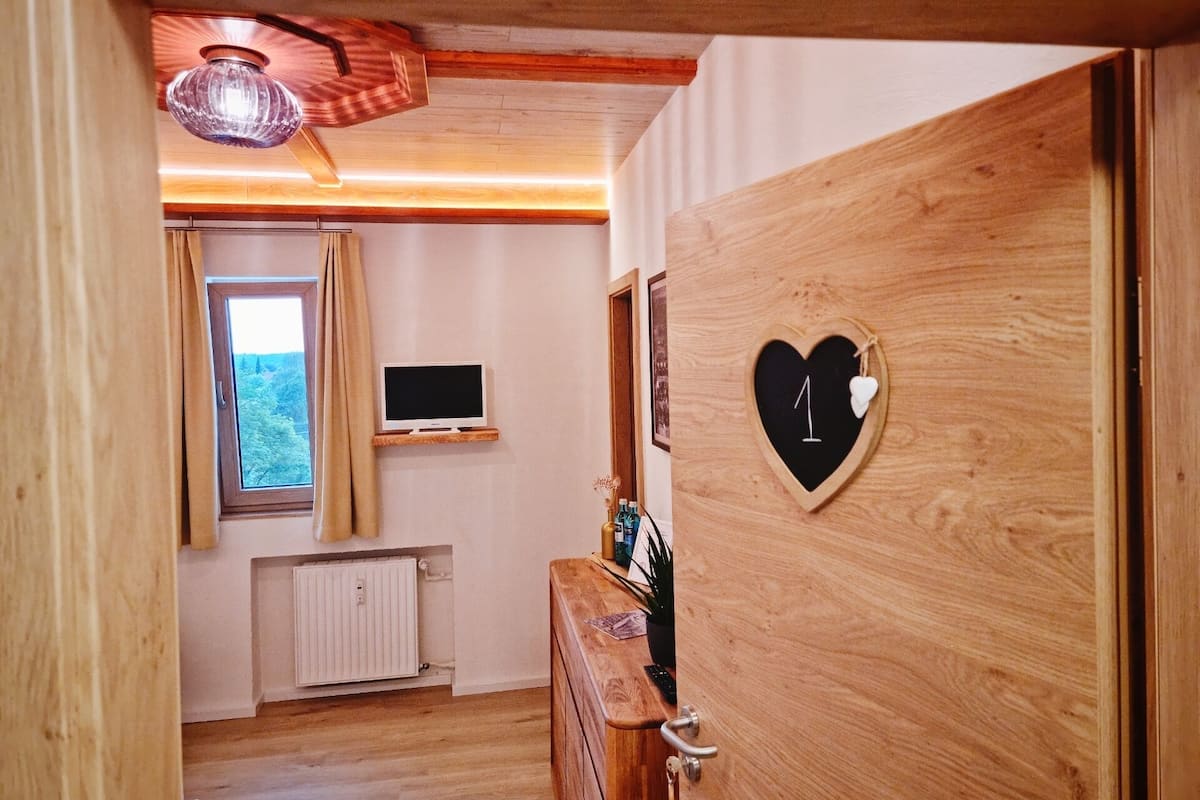 A wooden door with a heart-shaped decoration, a wooden dresser, a television on a shelf, and a window with curtains.