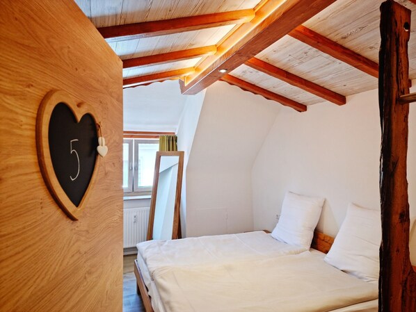 A cozy bedroom with a sloped ceiling, a wooden heart-shaped number '5' on the wall, a bed with white linens, and a window with curtains.