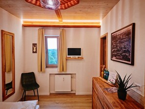 A room with a wooden ceiling, a window with curtains, a radiator, a television on a wooden shelf, a chair, a dresser with a plant, and a framed picture on the wall.
