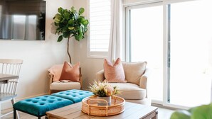Una sala de estar con sofá, reposapiés y mesa de centro, un televisor montado en la pared y un ventanal con persianas.