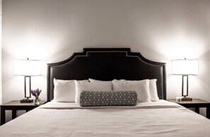 Ein ordentlich bezogenes Bett mit schwarzem Kopfteil, weißen Kissen und einem gemusterten Kissen. Auf dem Nachttisch befindet sich eine Lampe und ein Blumenstrauß.