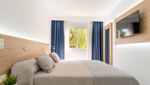 A modern bedroom with a bed, a TV mounted on the wall, a bedside table with a lamp, and a window with curtains. - Aparthotel Ciudad Laurel (Son Servera)