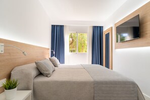A modern bedroom with a bed, a TV mounted on the wall, a bedside table with a lamp, and a window with curtains. - Aparthotel Ciudad Laurel (Son Servera)
