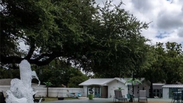 A swimming pool with a water fountain, surrounded by trees and a small building with outdoor seating. - 🧜‍♀️ Oasis Paradise Guesthouse (Granbury)