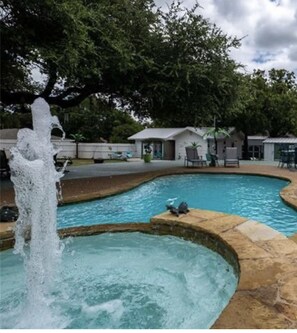 Piscina com fonte de água, cercada por árvores e um pequeno edifício com área de estar ao ar livre.