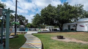A curved wooden pathway in a grassy area with a gazebo, a small pond, and a white building with a green roof. - 🧜‍♀️ Oasis Paradise Guesthouse (Granbury)