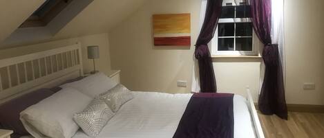 A bedroom with a bed, a nightstand, a lamp, a window with curtains, and a painting on the wall.