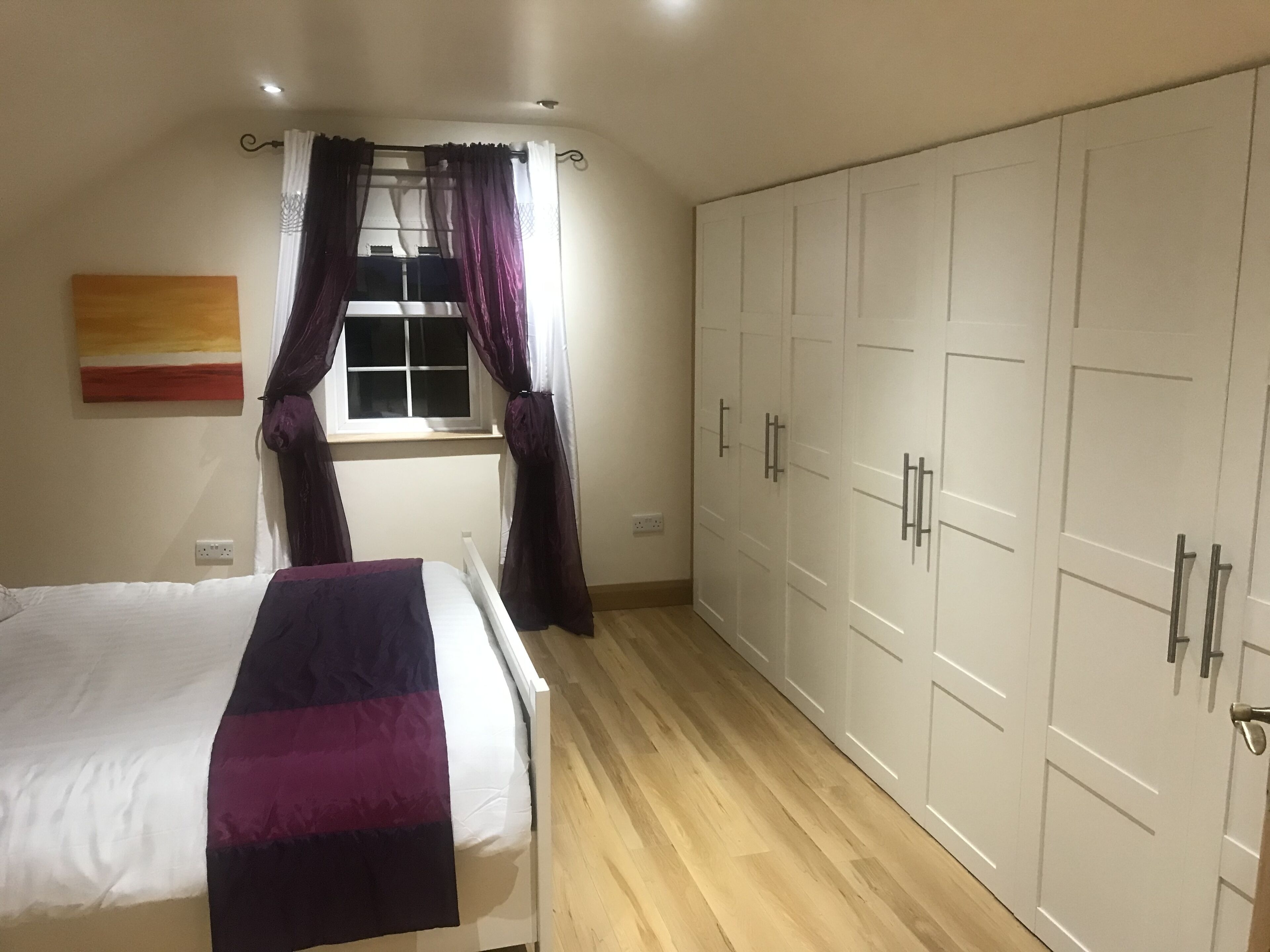 A bedroom with a bed, a window with curtains, a wardrobe, and a painting on the wall.