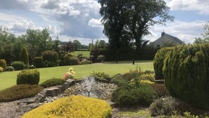 A well-maintained garden with a variety of shrubs and a lawn, a large tree, and a building in the background. - Dungannon 2 Bed Bungalow - Sleeps 5 - Pet Friendly (Dungannon)