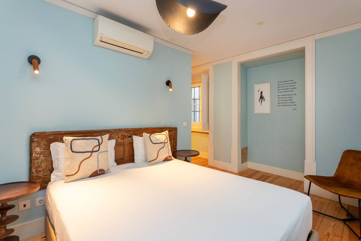 A bedroom with a large bed, an air conditioning unit, a wall-mounted light fixture, and a framed quote on the wall.
