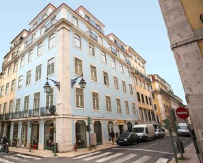 Un edificio de varios pisos con fachada azul claro, múltiples balcones y vista a la calle con coches y un paso de peatones.