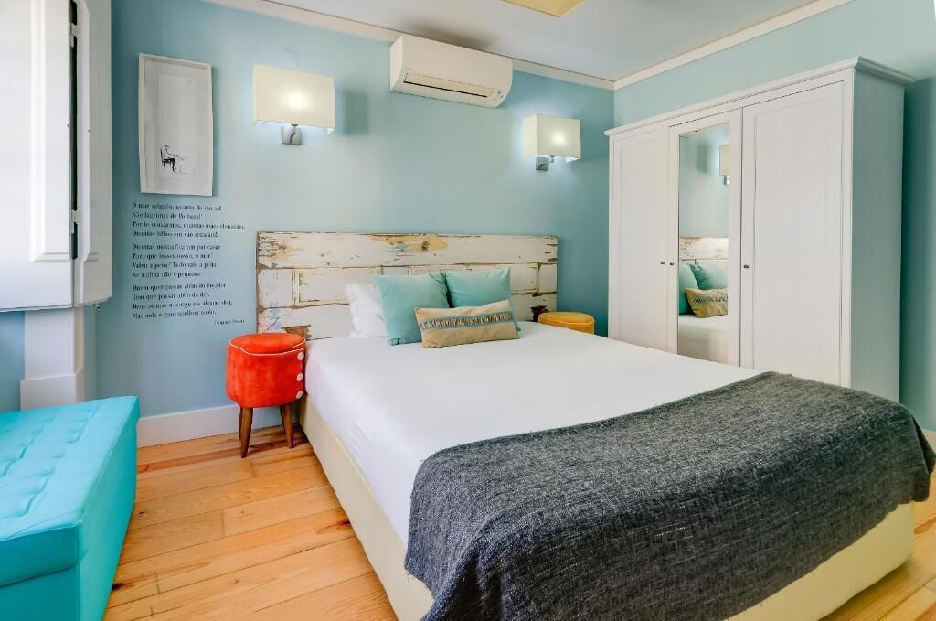 Een slaapkamer met een houten hoofdbord, een bed met een grijze deken, een rode kruk en een kledingkast.
