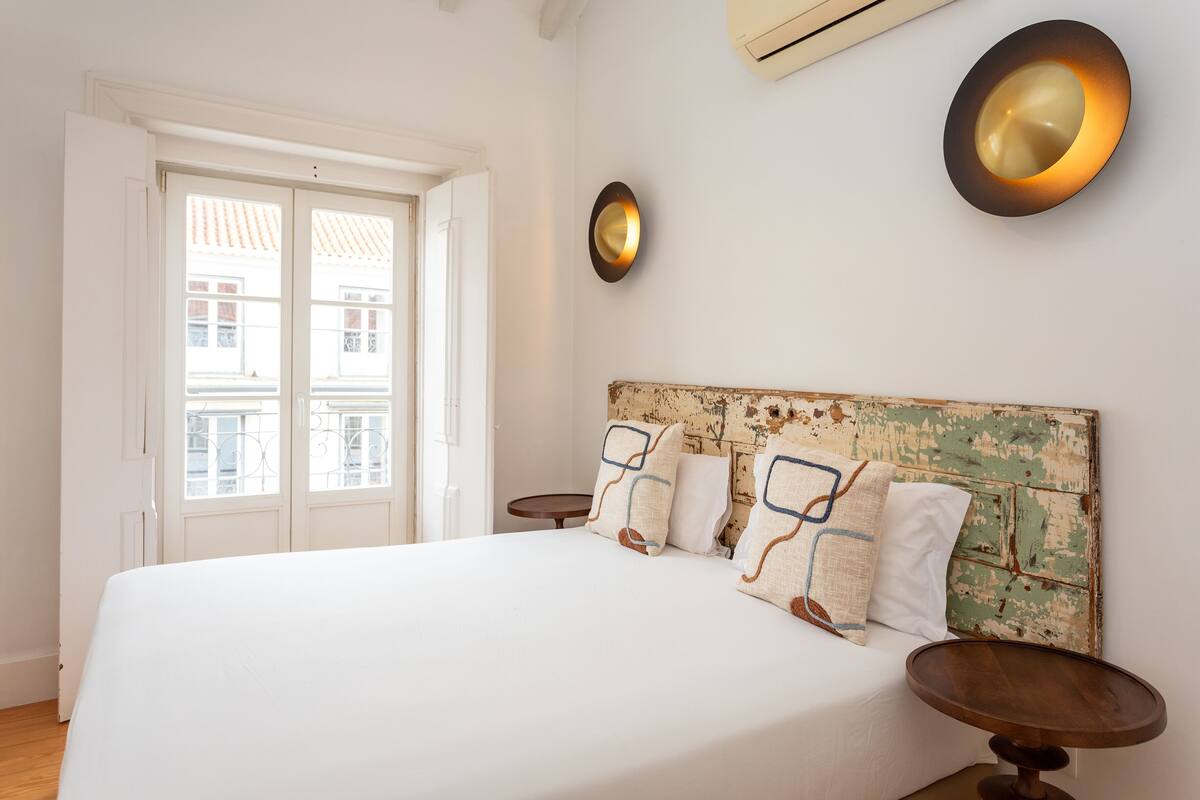 A bedroom with a large bed, a rustic headboard, bedside tables, and a window with a view of buildings.