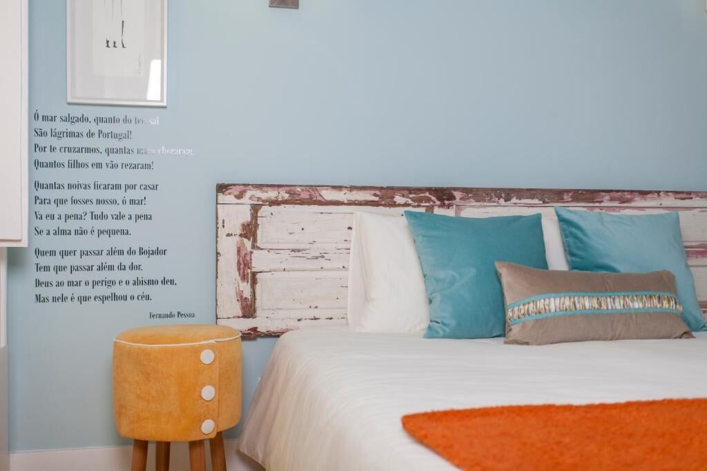 Ein Schlafzimmer mit einem weißen Bett, Kissen in Türkis und Grau, einem Holz-Kopfteil und einem kleinen Hocker.