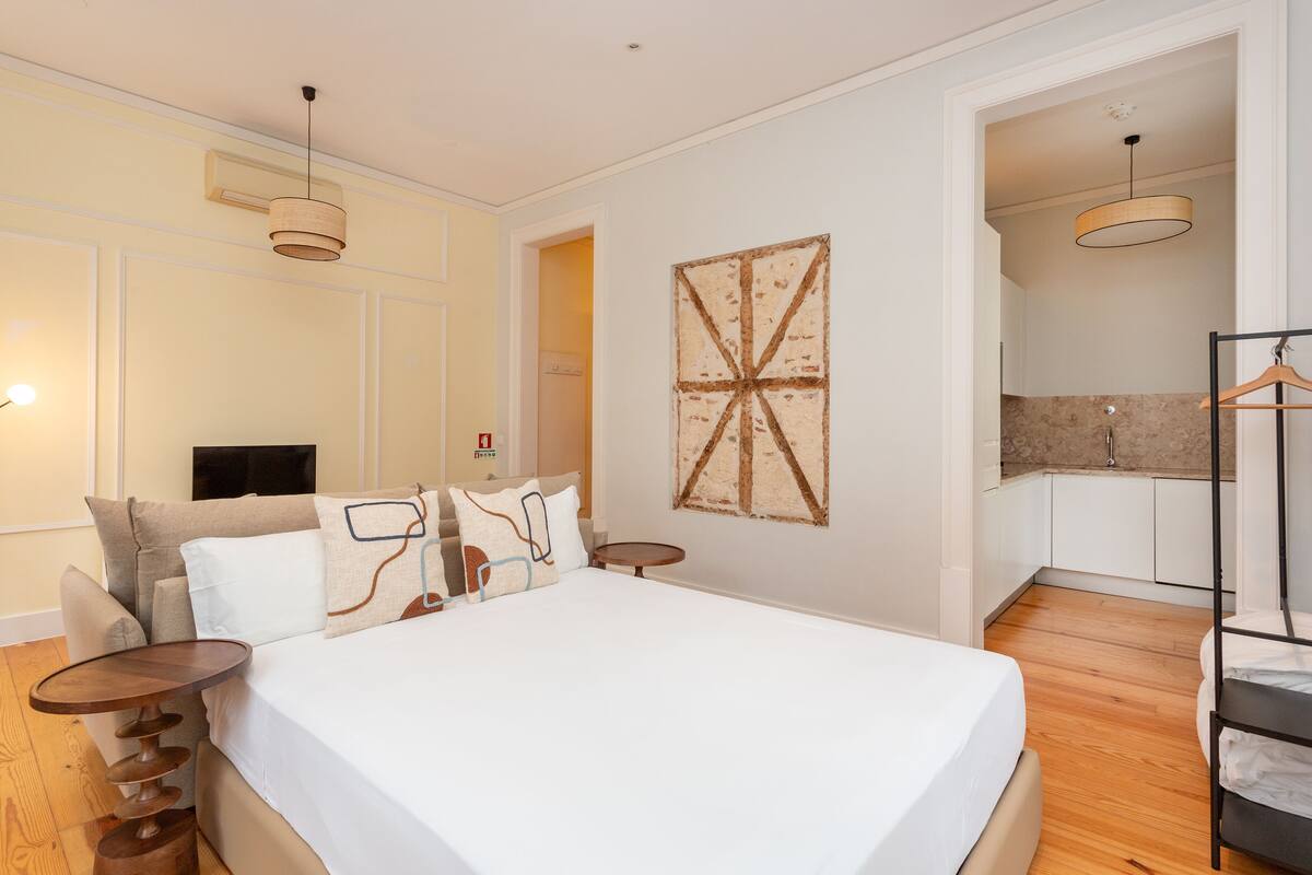 A bedroom with a large bed, bedside tables, a wall-mounted artwork, and a kitchen area visible through an open doorway.