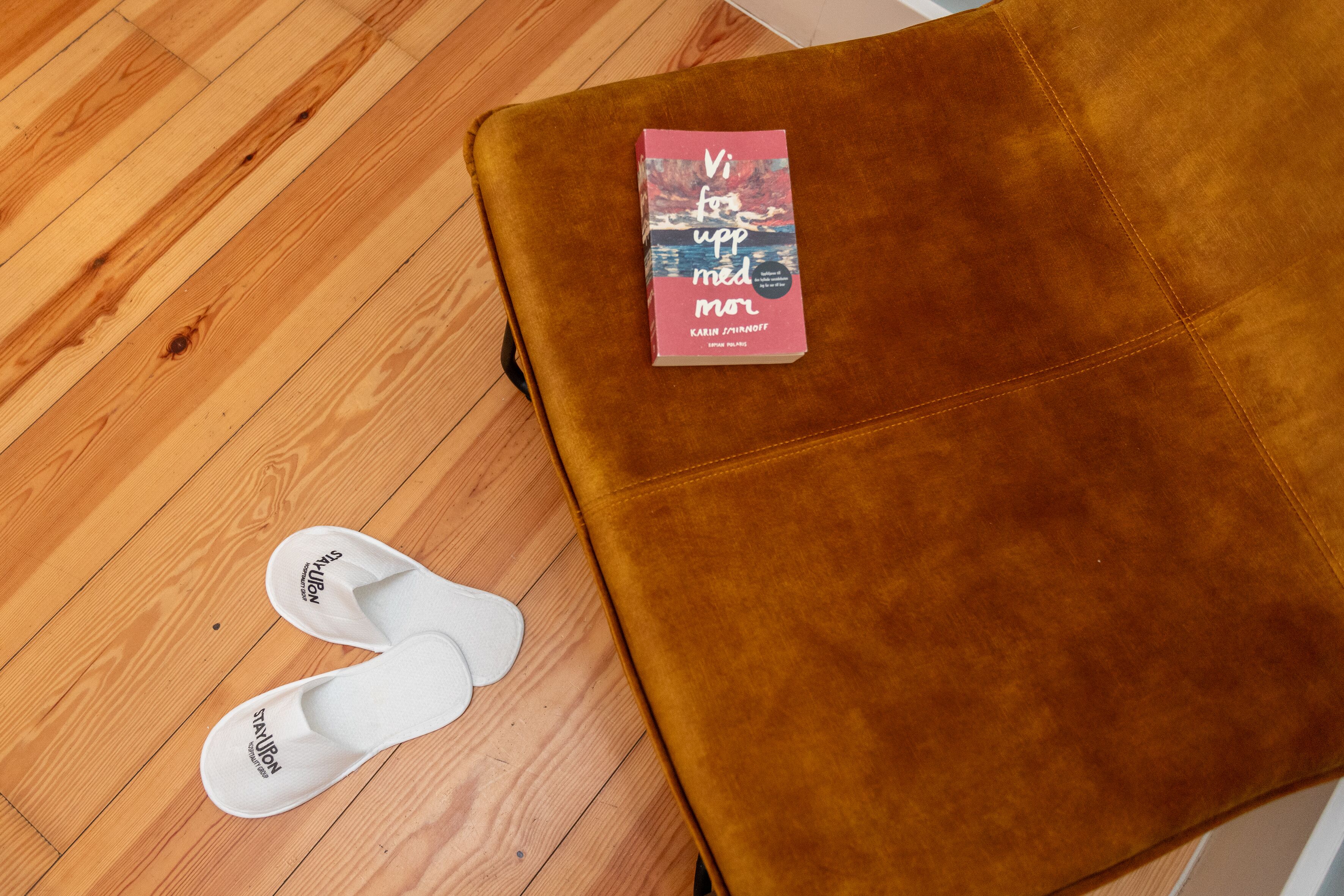 A pair of white slippers and a book titled "Vi for upp med mor" on a brown ottoman.
