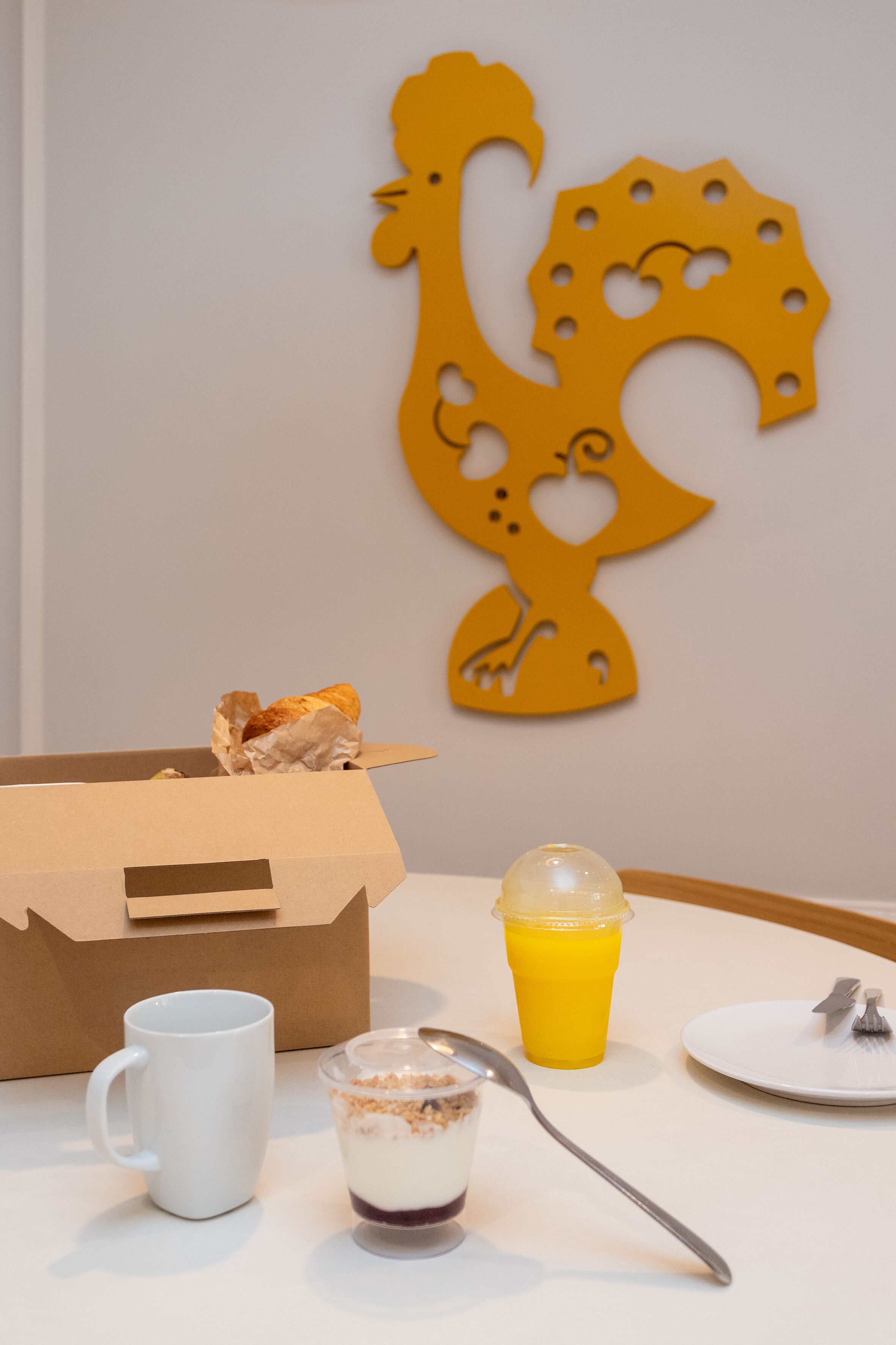 A breakfast setting with a cardboard box, a white mug, a glass of orange juice, a plate with a fork and spoon, and a dessert in a glass.